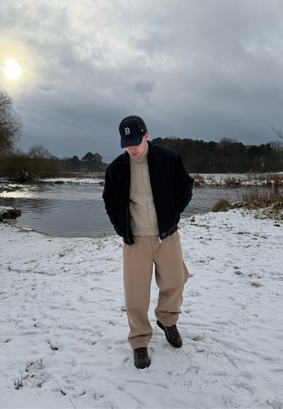 Hombre con chaqueta negra, suéter beige y pantalones caminando sobre un suelo cubierto de nieve cerca de un río bajo un cielo nublado.