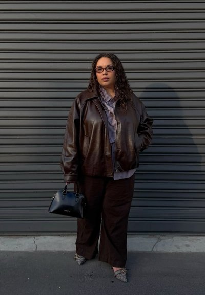 Mujer con cabello rizado y gafas, lleva chaqueta de cuero oscura, pantalones anchos, zapatos puntiagudos, sostiene un bolso negro, de pie junto a una persiana metálica gris.
