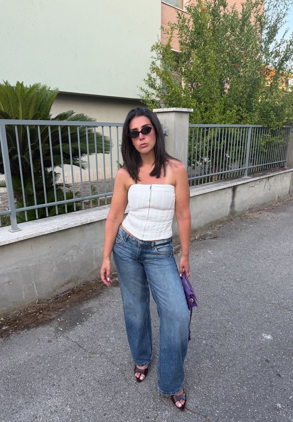 Femme aux cheveux foncés portant des lunettes de soleil noires, un haut blanc sans bretelles, un jean large et des talons, debout sur un trottoir tenant un petit sac à main violet.