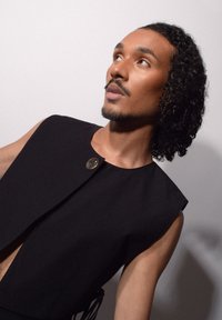 Svart ärmlös plagg med en rund knappdetalj, som visar upp en minimalistisk design och texturerad tyg, buren i en vinkel mot en neutral bakgrund.