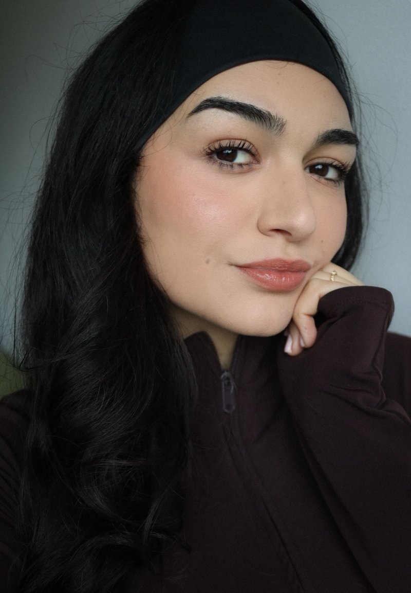 Jeune femme aux longs cheveux noirs portant un bandeau noir et un haut zippé foncé, reposant le menton sur la main avec un maquillage léger et une expression neutre.