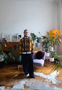 Γυναίκα με πλεκτό πουλόβερ με κίτρινες-καφέ ρόμβους και μπλε παντελόνι στέκεται σε δωμάτιο γεμάτο φυτά με μια λιλά καρέκλα, γάτα πάνω σε ντουλάπι και υφασμάτινο χαλί.