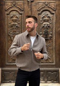 Braune Reißverschlussjacke aus weichem Stoff mit Kragen und gerippten Bündchen, getragen über einem weißen Hemd. Im Hintergrund sind verzierte Holztüren zu sehen.