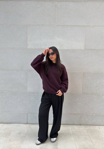 Sudadera burdeos de corte oversize, pantalones anchos negros y zapatos grises. Acompañados de gafas de sol negras estrechas frente a una pared de piedra.