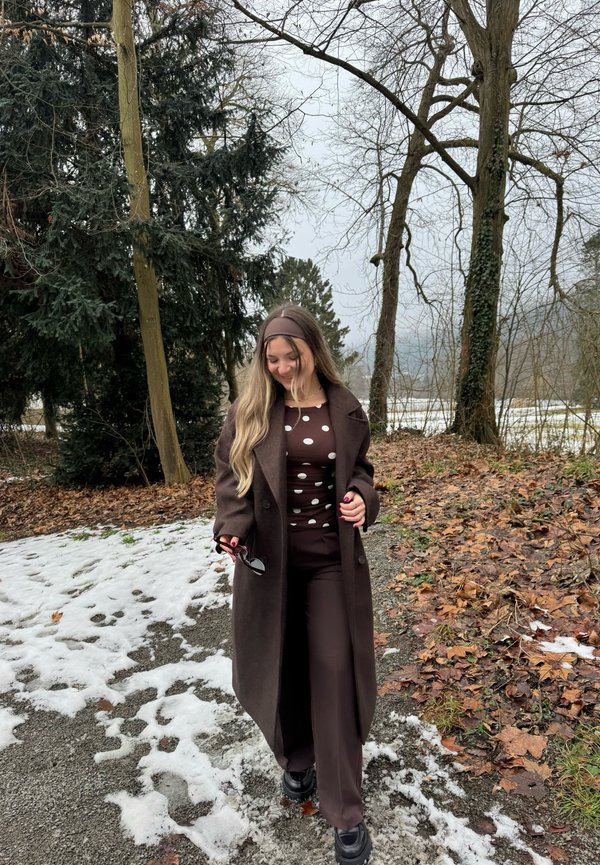 Femme marchant sur un chemin partiellement couvert de neige, portant un manteau marron et un haut à pois, tenant des lunettes de soleil, avec des arbres sans feuilles et une forêt en arrière-plan.