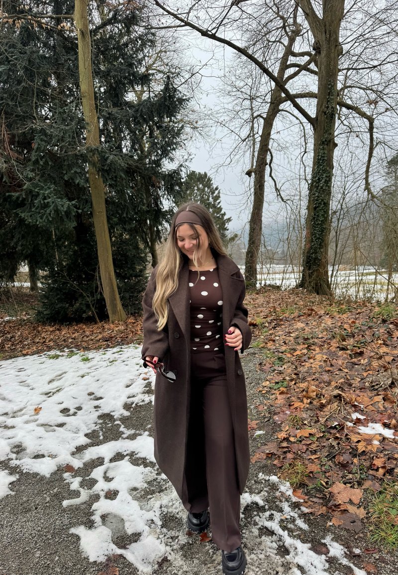 Femme marchant sur un chemin partiellement couvert de neige, portant un manteau marron et un haut à pois, tenant des lunettes de soleil, avec des arbres sans feuilles et une forêt en arrière-plan.