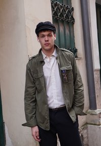 Olivegrüne Jacke über einem weißen Hemd, schwarze Mütze und marineblaue Hose. Runde Brille hängt aus der Jackentasche. Ohrstöpsel in den Ohren.