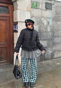 Personne portant une doudoune noire, un béret vert, des lunettes de soleil, un pantalon à carreaux, tenant un sac à main noir, debout près d'un mur en pierre avec un panneau de rue vert.