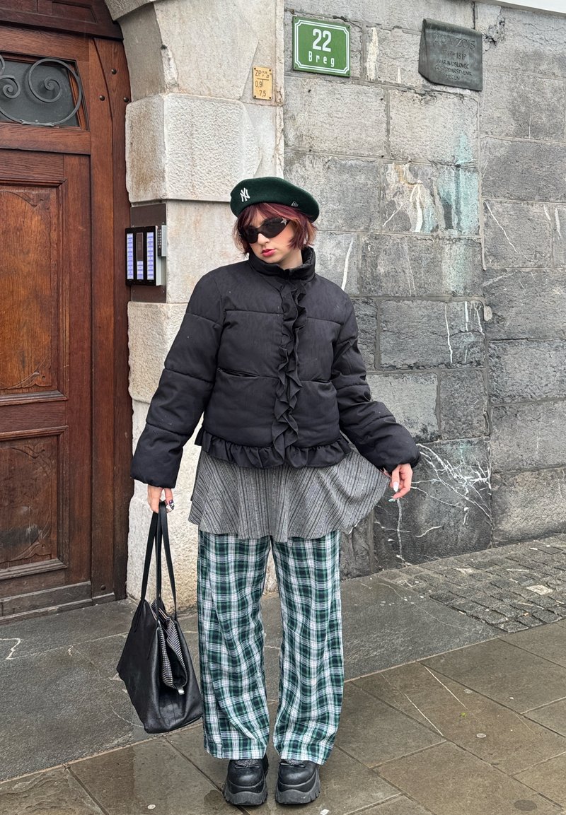 Person trägt eine schwarze Steppjacke, grünes Barett, Sonnenbrille, karierte Hose, hält eine schwarze Handtasche und steht an einer Steinstützmauer mit einem grünen Straßenschild.