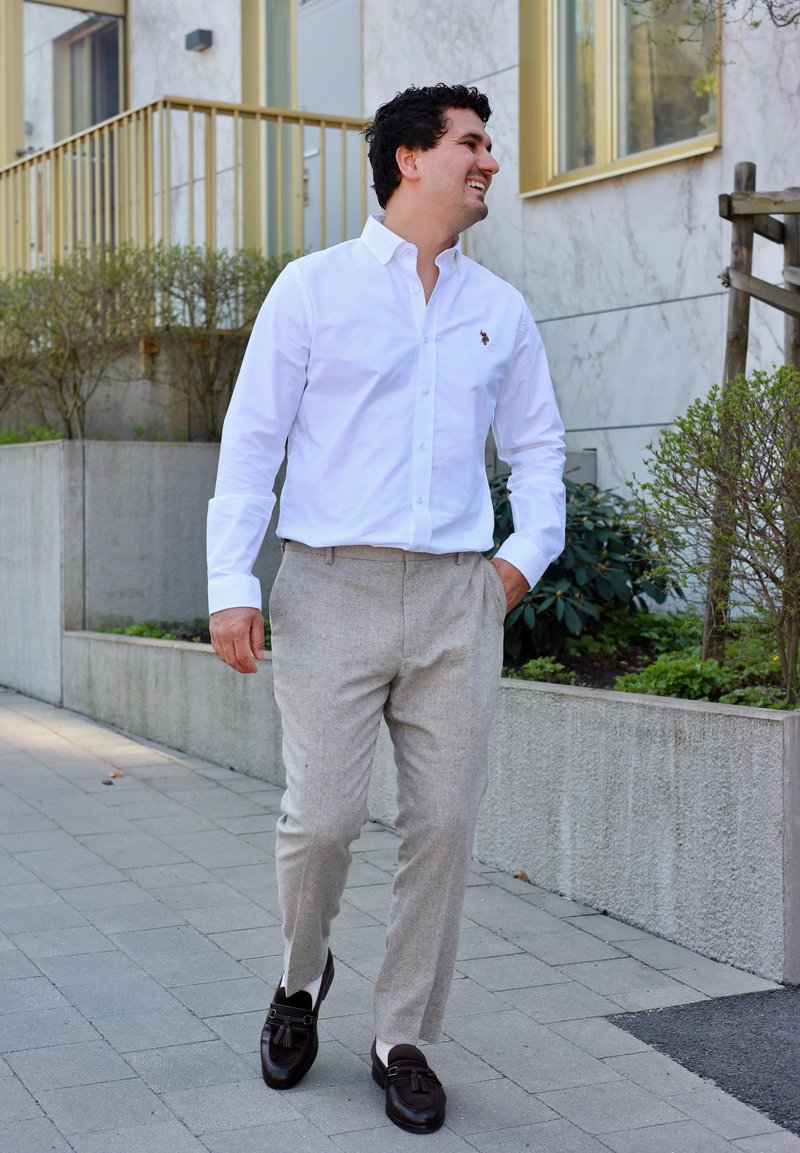 Homme en chemise blanche et pantalon beige marchant sur le trottoir urbain, une main dans la poche, souriant et regardant de côté.