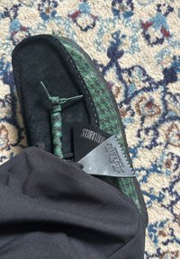 Fot iført grønn og svart tøysko med svart merkelapp, svarte bukser synlige, på mønstret krem, blå og rød teppebakgrunn.