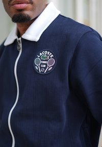 Giacca zip con cappuccio blu navy con colletto bianco, tessuto strutturato e logo ricamato del Lacoste Tennis Club con racchette e coccodrillo.