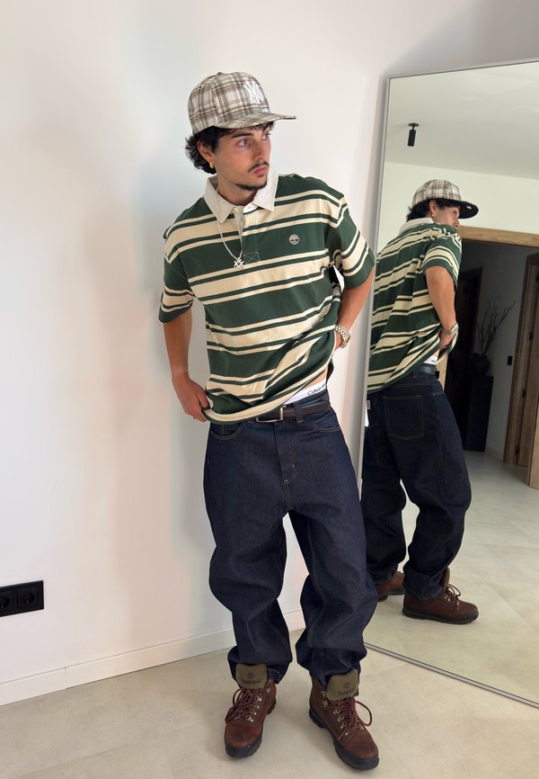 Jeune homme portant un polo rayé vert et crème, un jean sombre ample, des bottes marron et une casquette à carreaux, debout près d’un miroir à l’intérieur.