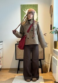 Jeune femme portant une veste marron à carreaux, un pantalon marron à larges rayures, un chapeau tressé et un sac à bandoulière rouge, debout à l'intérieur en tenant la tresse de son chapeau.