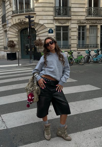 Joven mujer con gafas de sol, sudadera gris con capucha, pantalones cortos de cuero negro y zapatillas deportivas posando en un paso de peatones de la ciudad con bicicletas aparcadas y un edificio histórico detrás.