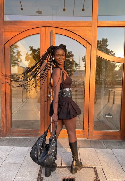 Vestido negro con dobladillo de volantes, peinado trenzado, botas negras hasta la rodilla y un bolso negro con detalles metálicos, posando junto a una puerta de madera.