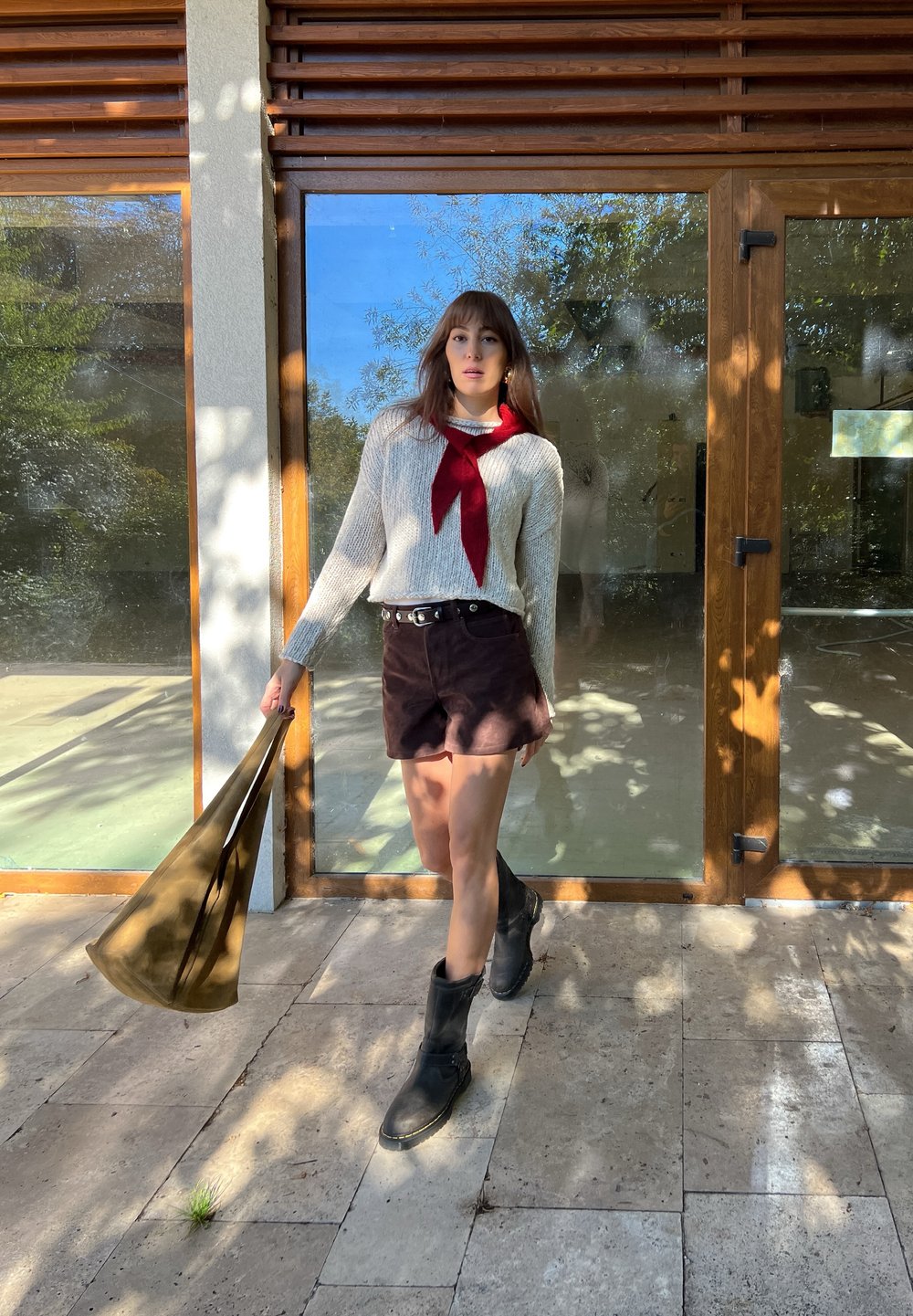 Crème gebreide trui met een rode lintdetail, bruine suède shorts, zwarte kniehoge laarzen en een groene totebag, staande bij een glazen deur.