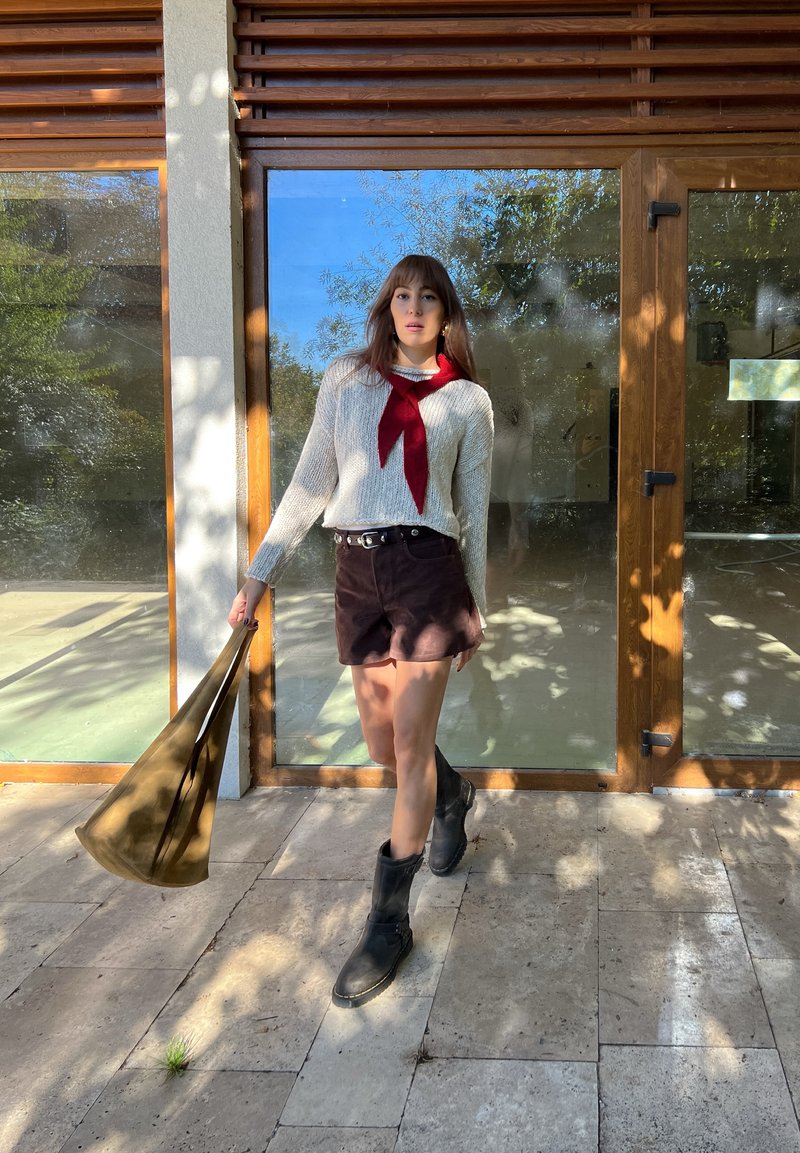 Kremfarget strikkegenser med rød bånddetalj, brune semskede shorts, svarte knelange støvler og en grønn totebag, stående ved en glassdør.