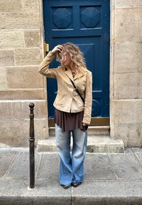 Vrouw met krullend haar in een beige jas, bruine top, wijde jeans en een crossbody tas staat op de stoep naast een blauwe deur.