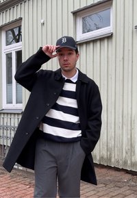 Jeune homme portant un manteau noir, un pull rayé, un pantalon gris et une casquette de baseball noire, debout à l'extérieur près d'un bâtiment de couleur claire, touchant la visière de sa casquette.
