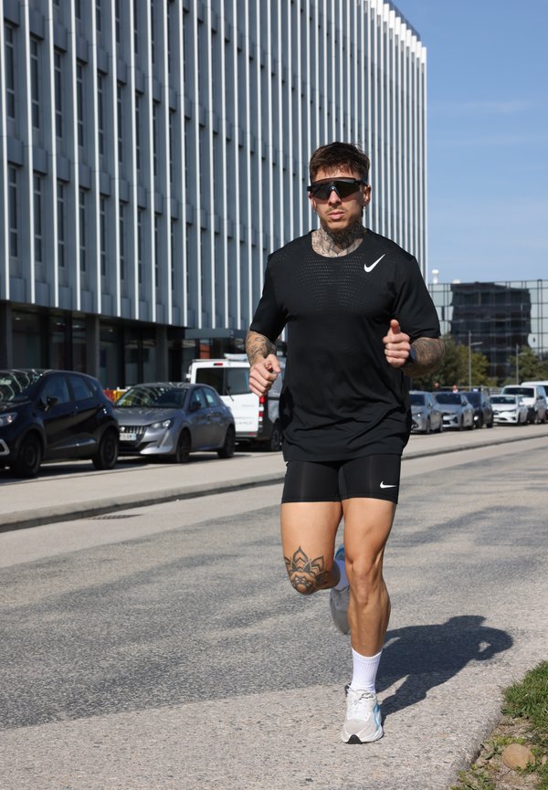 Homme tatoué portant une tenue de sport noire et des lunettes de soleil en train de jogger dans une rue de la ville avec des voitures garées et des bâtiments modernes en arrière-plan.