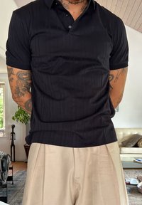 Man met getatoeëerde armen, gekleed in een zwart poloshirt en beige broek, staat in een woonkamer met planten en meubels op de achtergrond.