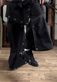 Pantalon noir à jambes larges avec des boutons-pression argentés, associé à des baskets noires présentant des détails texturés. Fond en bois avec accent vert.