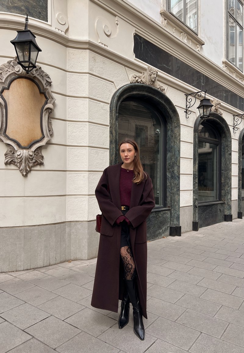 Manteau en laine bordeaux avec un large col, associé à un pull bordeaux et une jupe noire. Portez des bottes noires montant jusqu'aux genoux et des collants en dentelle.