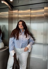 Camicia a righe blu chiaro con colletto bianco oversize abbinata a pantaloni bianchi larghi. Borsa intrecciata marrone scuro in mano.