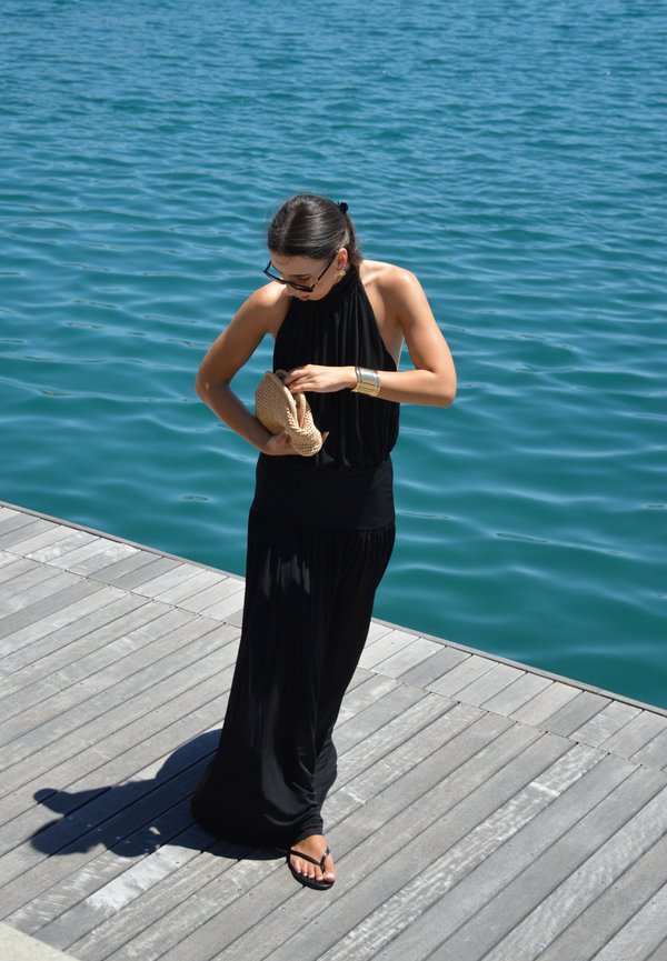Femme en robe noire à col halter et sandales, debout sur un quai en bois près de l'eau bleue, tenant un chapeau tressé beige et regardant vers le bas.