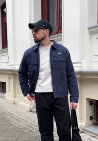 Veste bleu marine au design structuré, avec deux poches avant, portée sur un sweat-shirt blanc à col rond et un pantalon noir, accessoirisée d'une casquette noire et de lunettes de soleil.