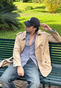 Man in a beige trench coat, blue checkered shirt, and black cap sitting on a green park bench, looking to the left, with greenery in the background.