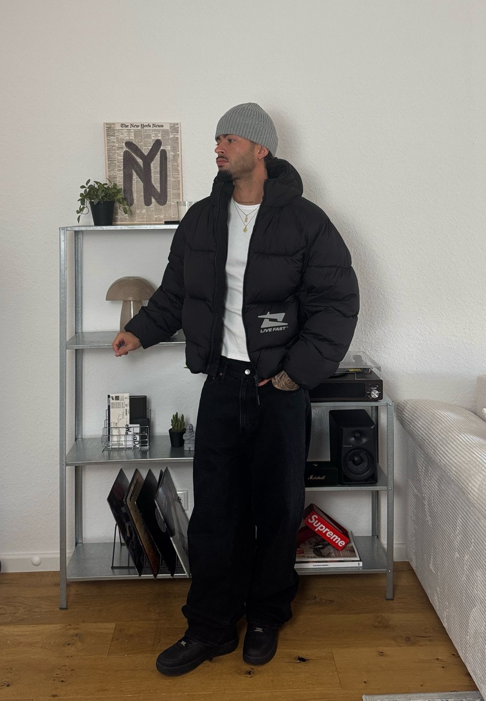 Mees, kellel on seljas hall beanie ja must puffer-jakk, seisab metallriigi kõrval, kus on vinüülplaadid, kõlarid ja kaunistused minimalistlikus elutoas.