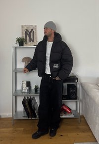 Mees, kellel on seljas hall beanie ja must puffer-jakk, seisab metallriigi kõrval, kus on vinüülplaadid, kõlarid ja kaunistused minimalistlikus elutoas.