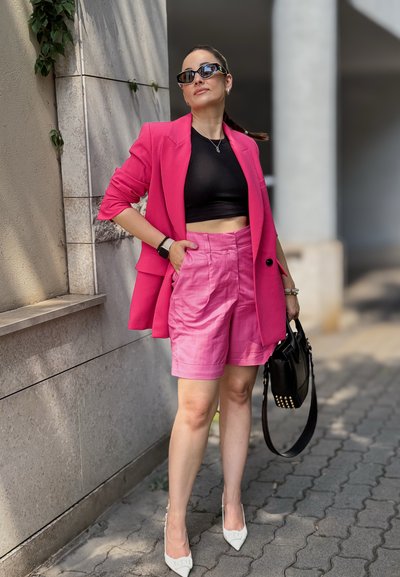 Blazer rosa brillante sobre un top corto negro, combinado con pantalones cortos rosa claro. Tacones blancos de punta y un bolso negro con tachuelas completan el look.