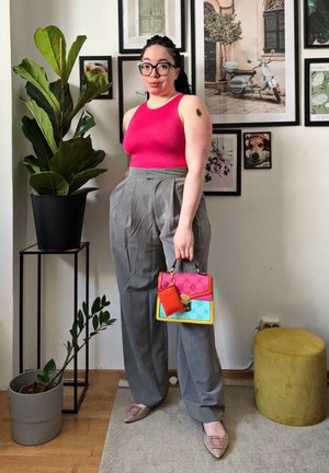 Pantalones de cintura alta grises, top de tirantes fucsia, bolso rosa, azul y amarillo con un acento rojo. Bailarinas grises con una hebilla decorativa. Fondo de planta verde.