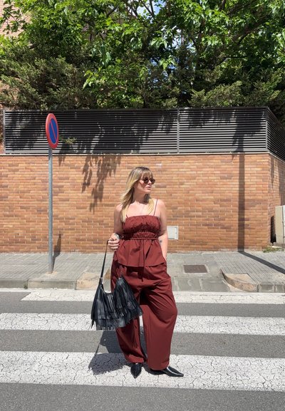 Mujer con top sin mangas rojo y pantalones, llevando un bolso negro con flecos, de pie en un paso de peatones junto a una pared de ladrillos y una señal de prohibido aparcar.