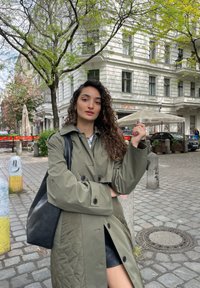 Cappotto trapuntato verde oliva con grandi bottoni neri, indossato sopra una mini gonna nera. La persona porta una borsa a tracolla nera. Sfondo di una strada di ciottoli.