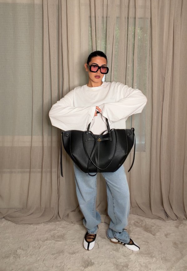 Personne portant une chemise blanche à manches longues oversize, un jean bleu, de grandes lunettes de soleil foncées, tenant un grand sac à main noir, debout sur un tapis beige.