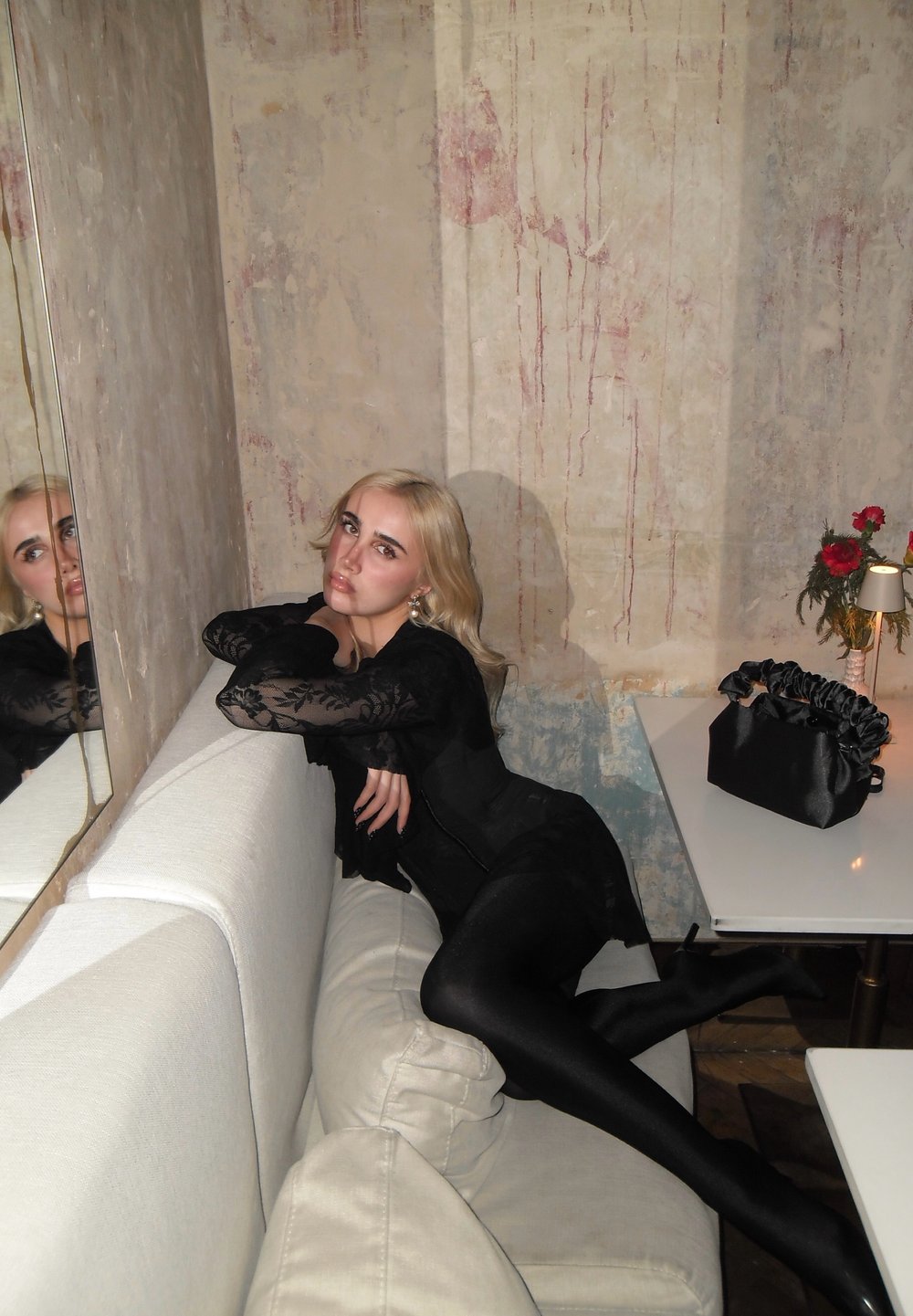 Jeune femme aux cheveux blonds en robe en dentelle noire s'allongeant sur un canapé crème, près d'un mur usé et d'une table blanche avec un sac noir et une petite lampe.