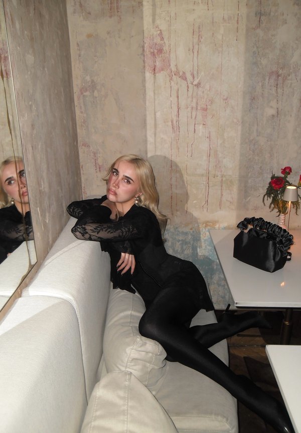 Ung kvinne med blondt hår i svart blonderkjole som lener seg tilbake på kremfarget sofa, nær en slitt vegg og et hvitt bord med svart veske og liten lampe.