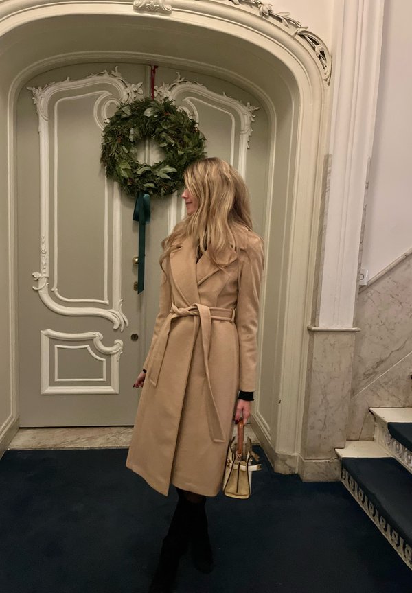 Abrigo envolvente de lana beige con cintura ajustable, cuello de muesca y mangas largas. De pie frente a una puerta decorada con una corona verde.