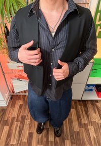 Pessoa vestindo uma camisa preta de riscas finas, colete preto, jeans largos azuis e sapatos castanhos escuros brilhantes, em pé sobre um chão de madeira no interior.