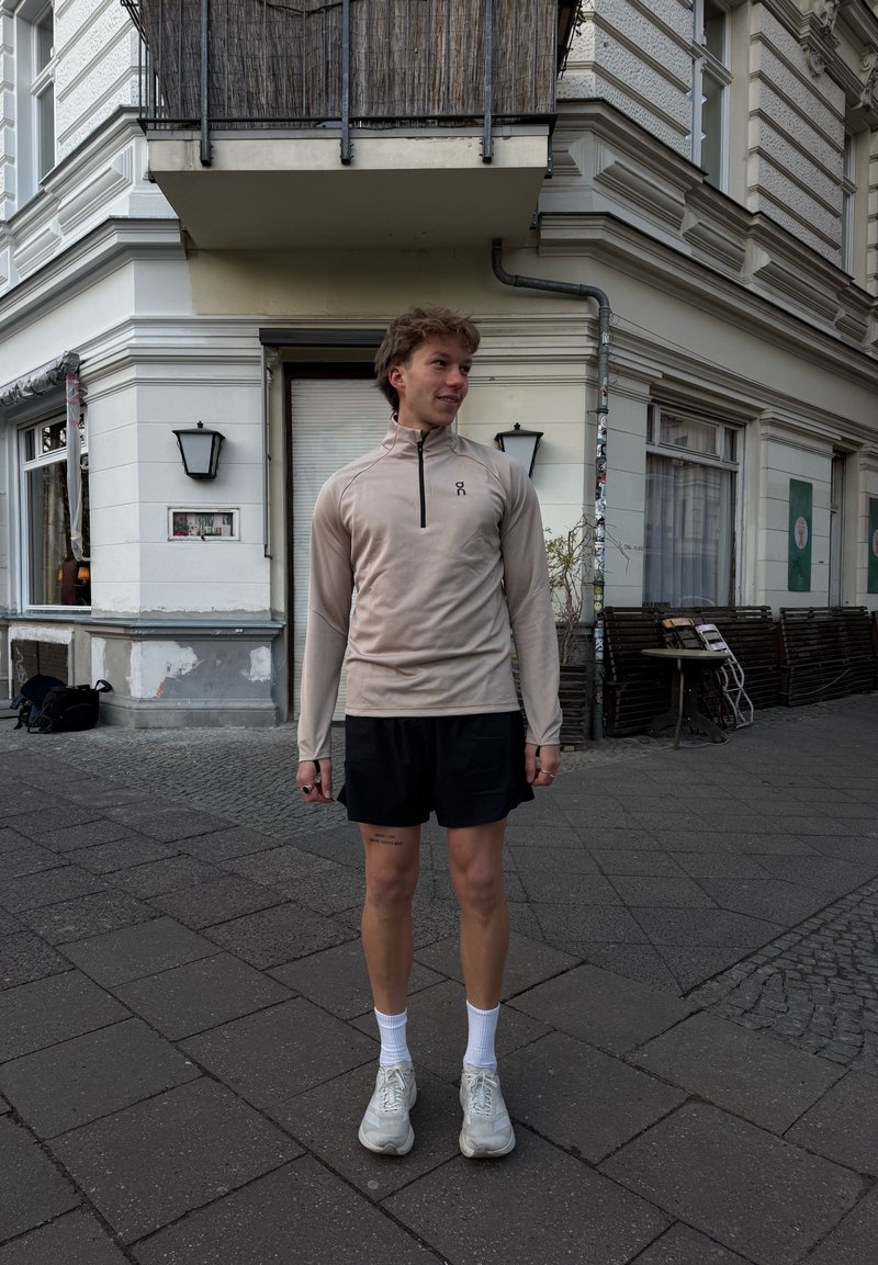 Junger Mann in beigefarbenem Langarmshirt und schwarzen Shorts steht auf einem Bürgersteig der Stadt in der Nähe eines Gebäudes mit Außensitzbereich.