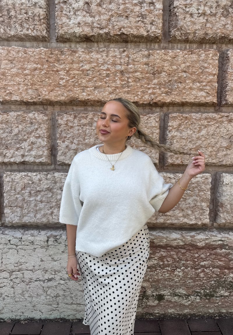 Jeune femme aux yeux fermés tenant ses cheveux tressés, vêtue d’un pull blanc et d’une jupe blanche à motifs noirs, debout contre un mur en pierre.