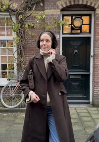 Palton maro cu dublu rând de nasturi mari, pulover crem cu guler înalt și căști auriculare închise la culoare. Blugi bleu deschis și o geantă mică maro.