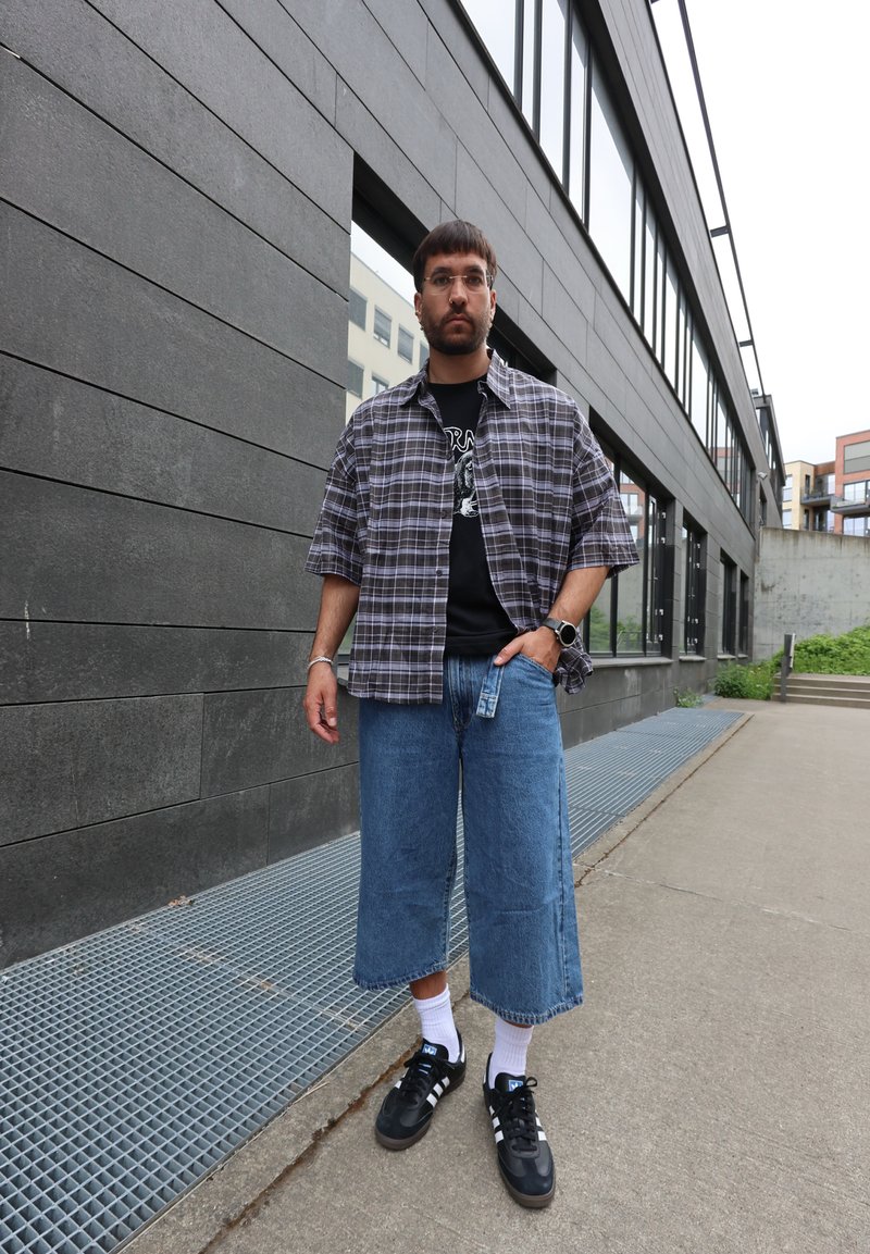 Homme portant une chemise à manches courtes à carreaux, un t-shirt noir graphique, un jean large coupé, des chaussettes blanches et des baskets noires, debout sur un trottoir en ville.