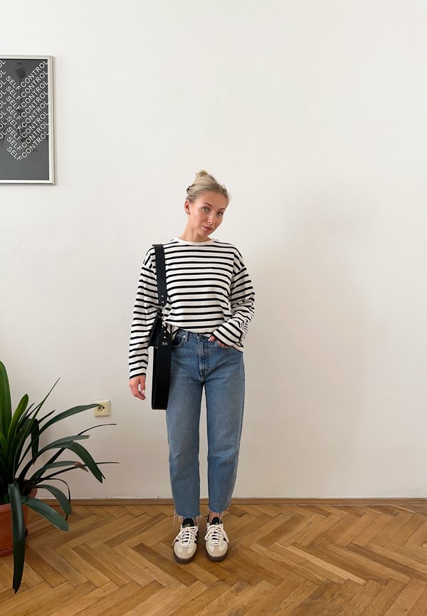 Camisola de mangas longas às riscas em preto e branco, jeans azuis e ténis brancos. Saco preto com alça. Piso de madeira e planta de interior ao fundo.