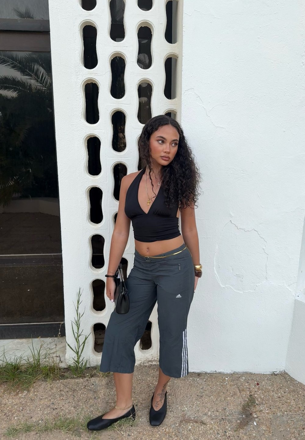 Top corto negro con cuello halter, pantalones cortos grises con tres franjas blancas, zapatos planos negros, combinado con un bolso pequeño y acentos dorados.