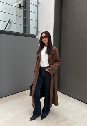 Mujer con abrigo largo marrón, camisa blanca, vaqueros oscuros, botas negras puntiagudas y gafas de sol, de pie contra una pared gris en el interior.